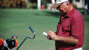 An elderly man in a red shirt and white cap stands on a golf course, holding a scorecard filled with birdies and pars near a cart with golf clubs.